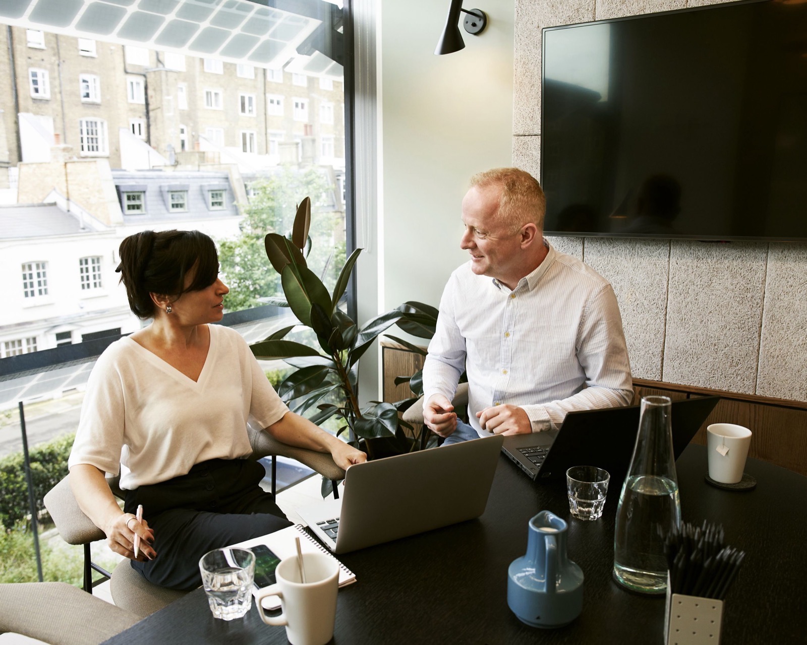 Two colleagues mid-conversation at a desk in a London office, terraced houses through the window