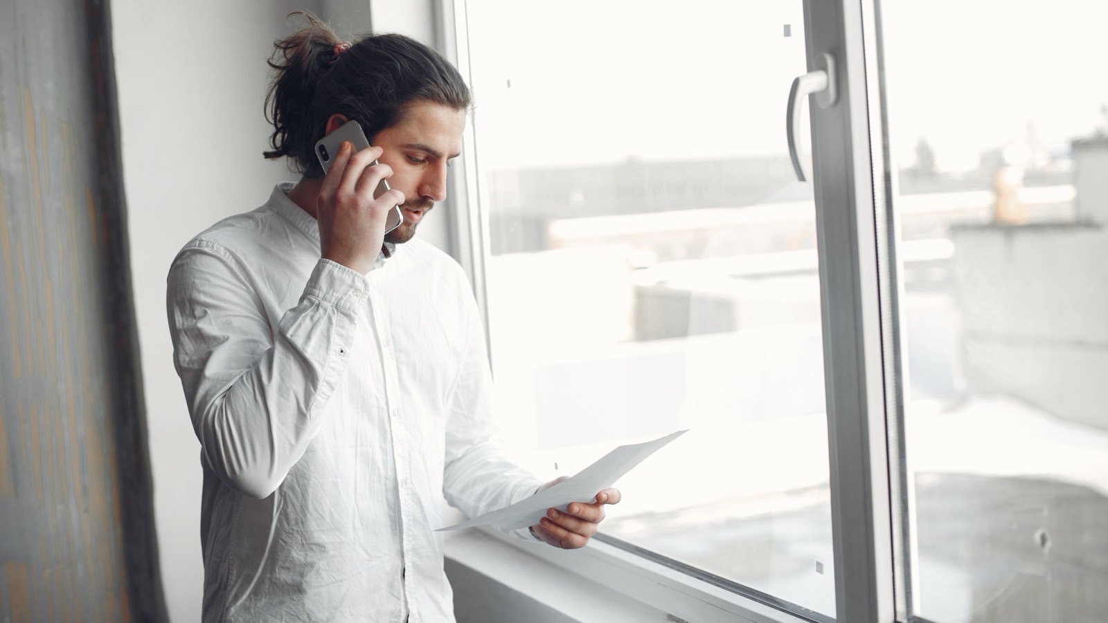 Someone on the phone at an office window, looking over a printed sheet